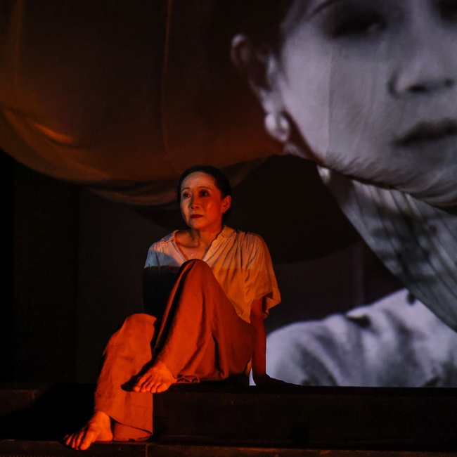 Woman lit in red lighting in front of a screen with a woman's face projected onto it