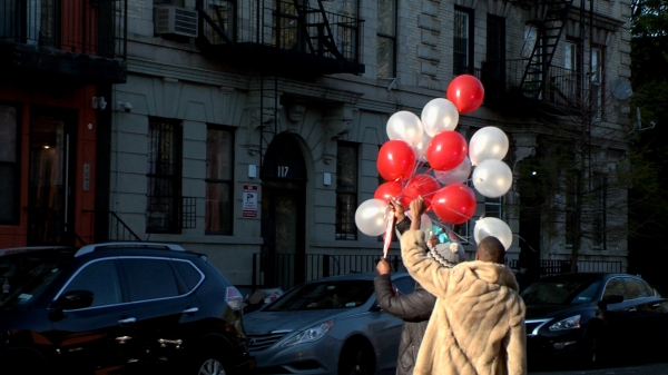 Two people holding red and white balloons in the air on a street corner with cars in the background
