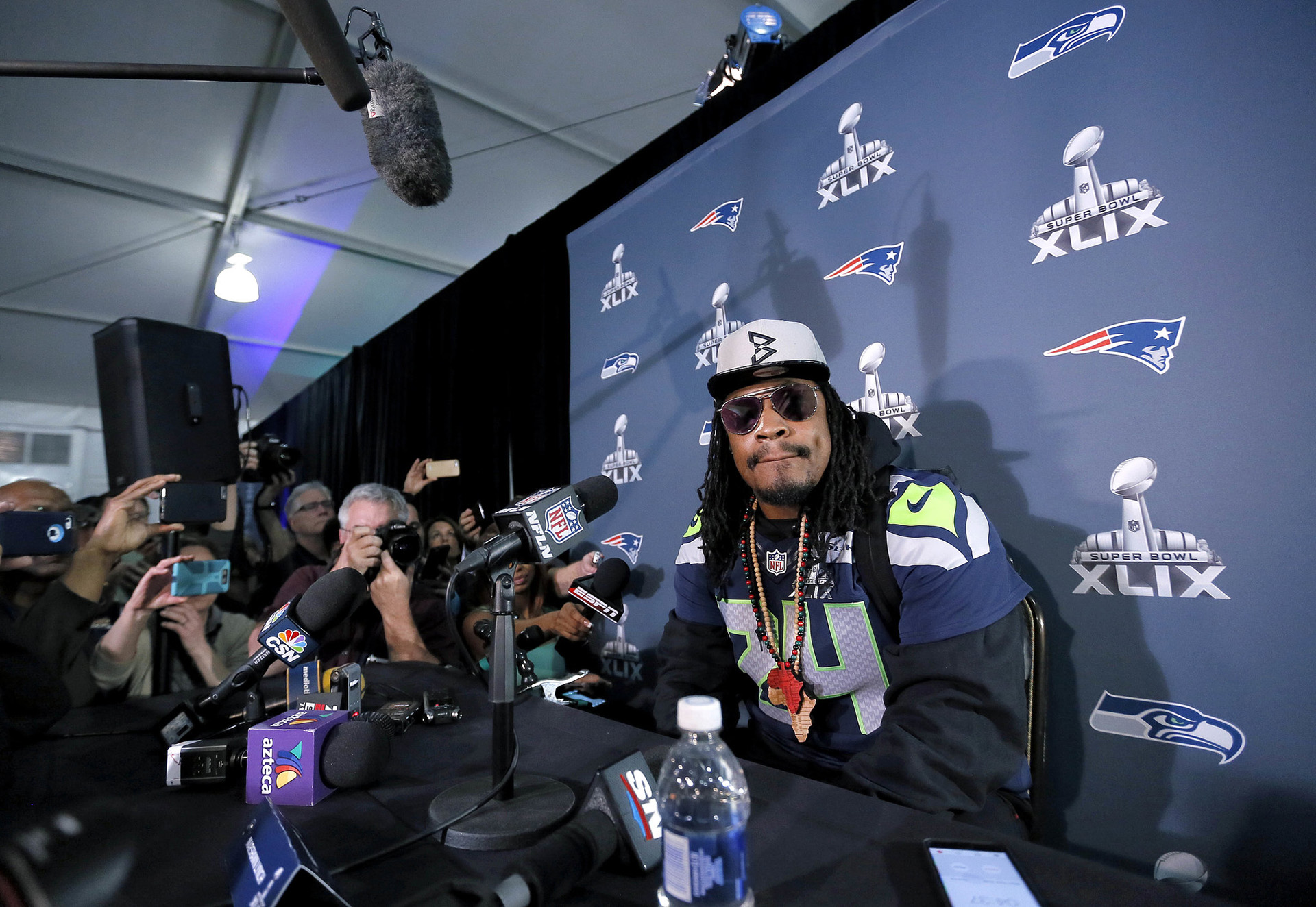 Marshawn Lynch sitting at a table surrounded by microphones