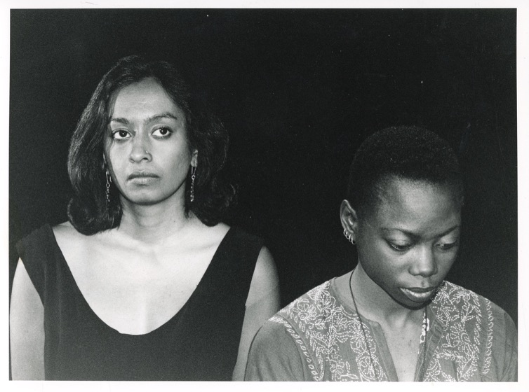 Two people stnading against a black background looking away from the camera
