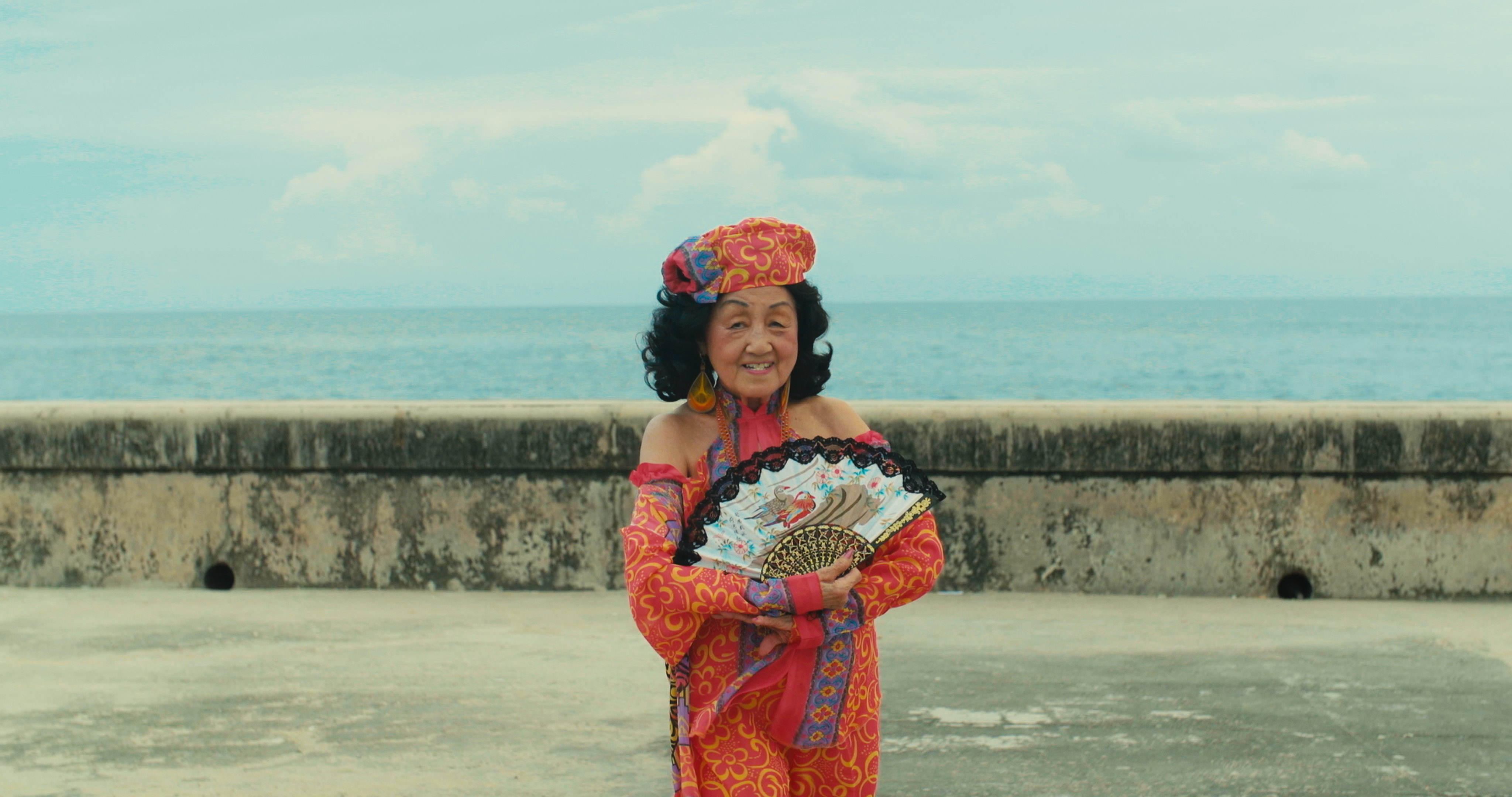 Woman standing in front of the ocean with a smile and holding a decorative fan.