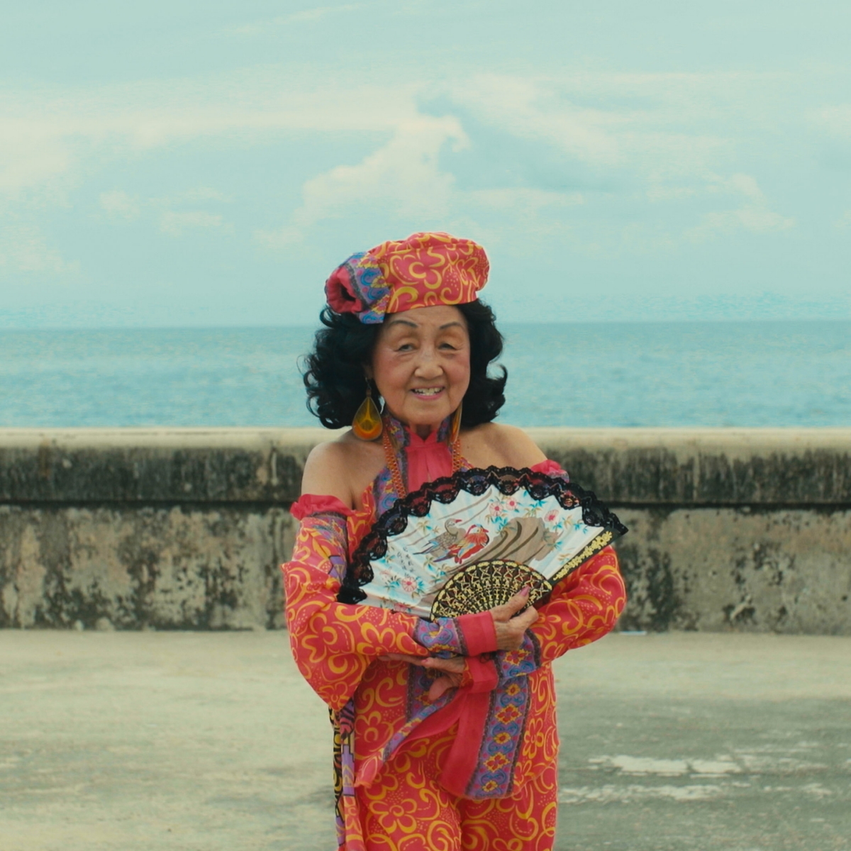 Woman standing in front of the ocean with a smile and holding a decorative fan.