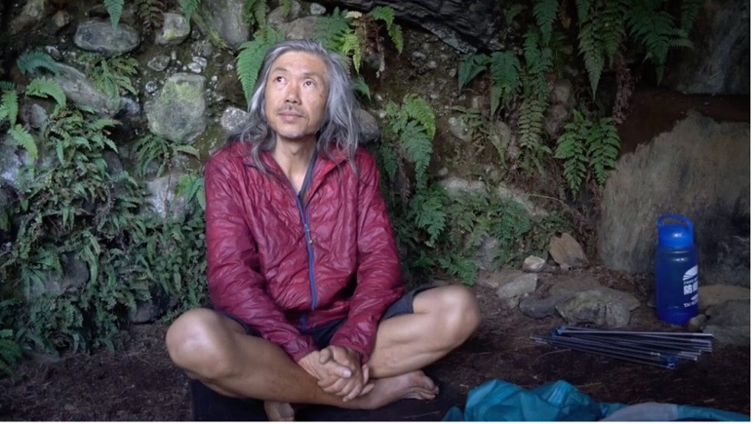 Man sitting cross-legged on the ground. He is wearing a red zip up jacket.