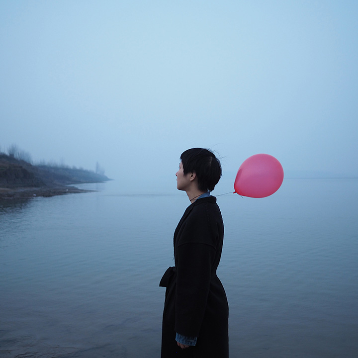 Woman with short hair facing left, wearing black coat and holding a red balloon.