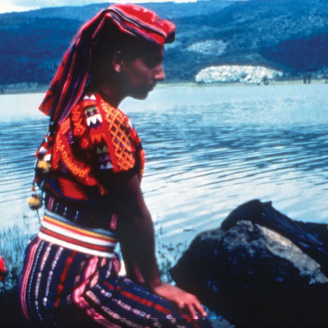 A woman in a brightly colored shirt kneeling on the shore of a body of water.