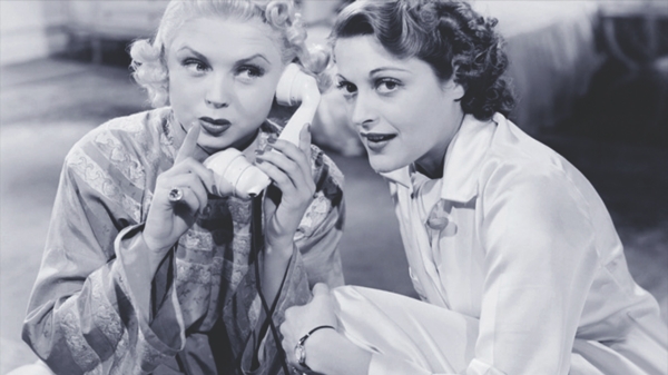 Two women sitting on the floor of a bedroom. One holding a hand to her lips and the phone in the other to her ear. The other is leaning towards the receiver.