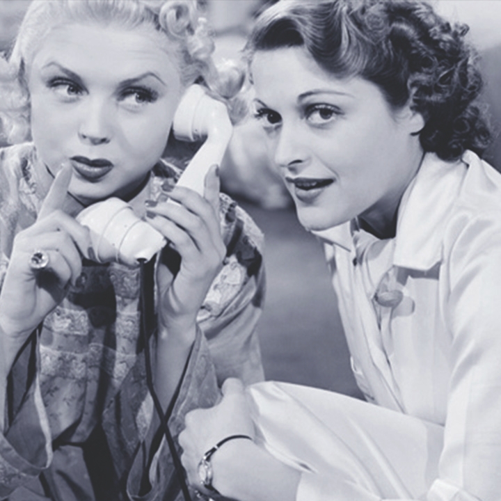 Two women sitting on the floor of a bedroom. One holding a hand to her lips and the phone in the other to her ear. The other is leaning towards the receiver.