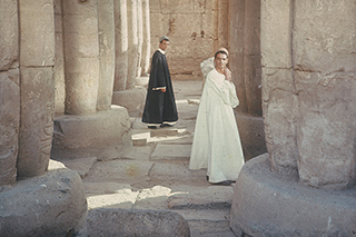Two figures standing between stone pillars, one in the background wearing black, the one in the foreground wearing white