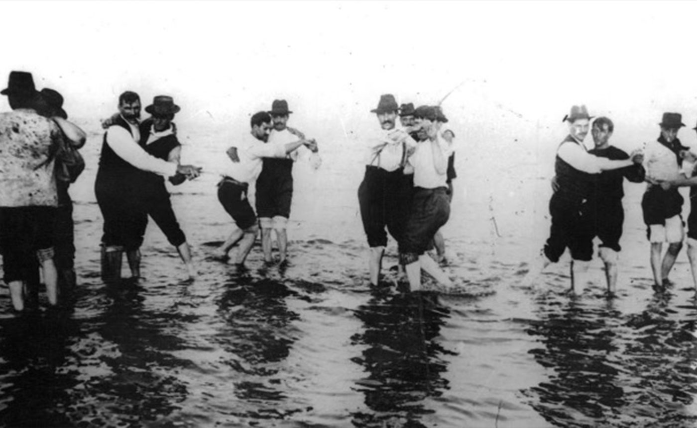 Black and white image of men in hats dancing together in shallow water