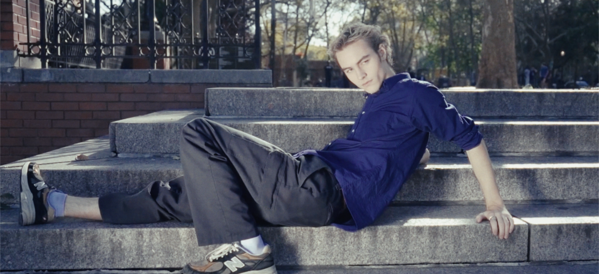 Cadel Martel sits on concrete steps in a park, one leg bent and the other stretched out on the step. He gazes off screen