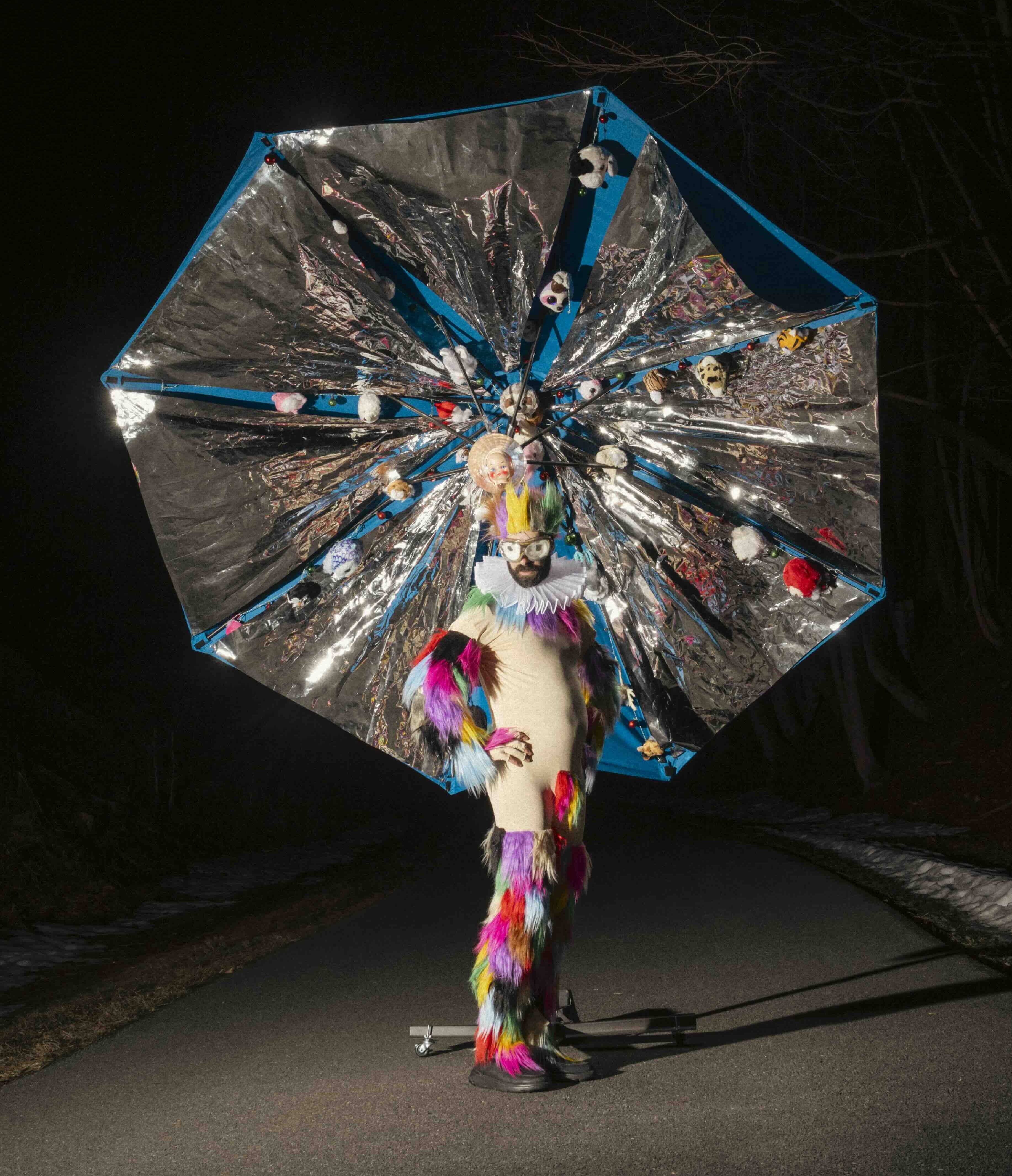 A figure in a costume with rainbow furs with a massive metallic umbrella behind them