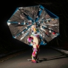 A figure in a costume with rainbow furs with a massive metallic umbrella behind them