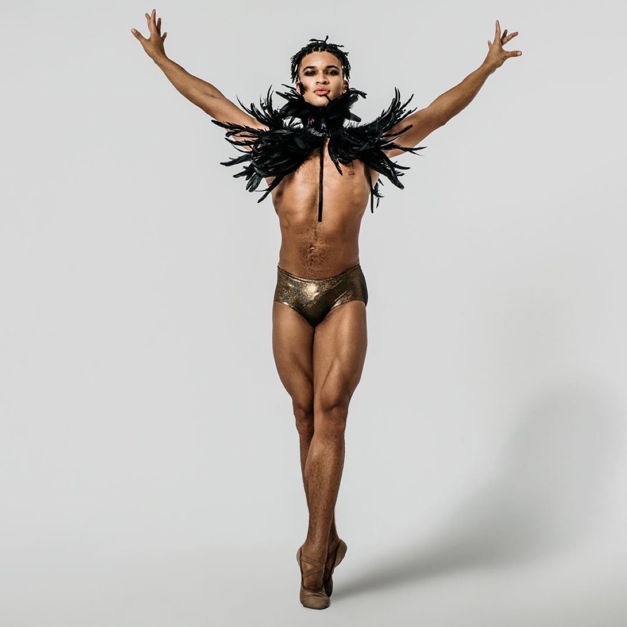 Dancer in white background wearing black feathers on shoulders and wearing ballet shoes.
