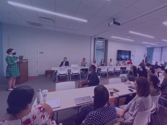 A facilitator stands to the left of the image. A classroom full of diverse individuals listens intently.