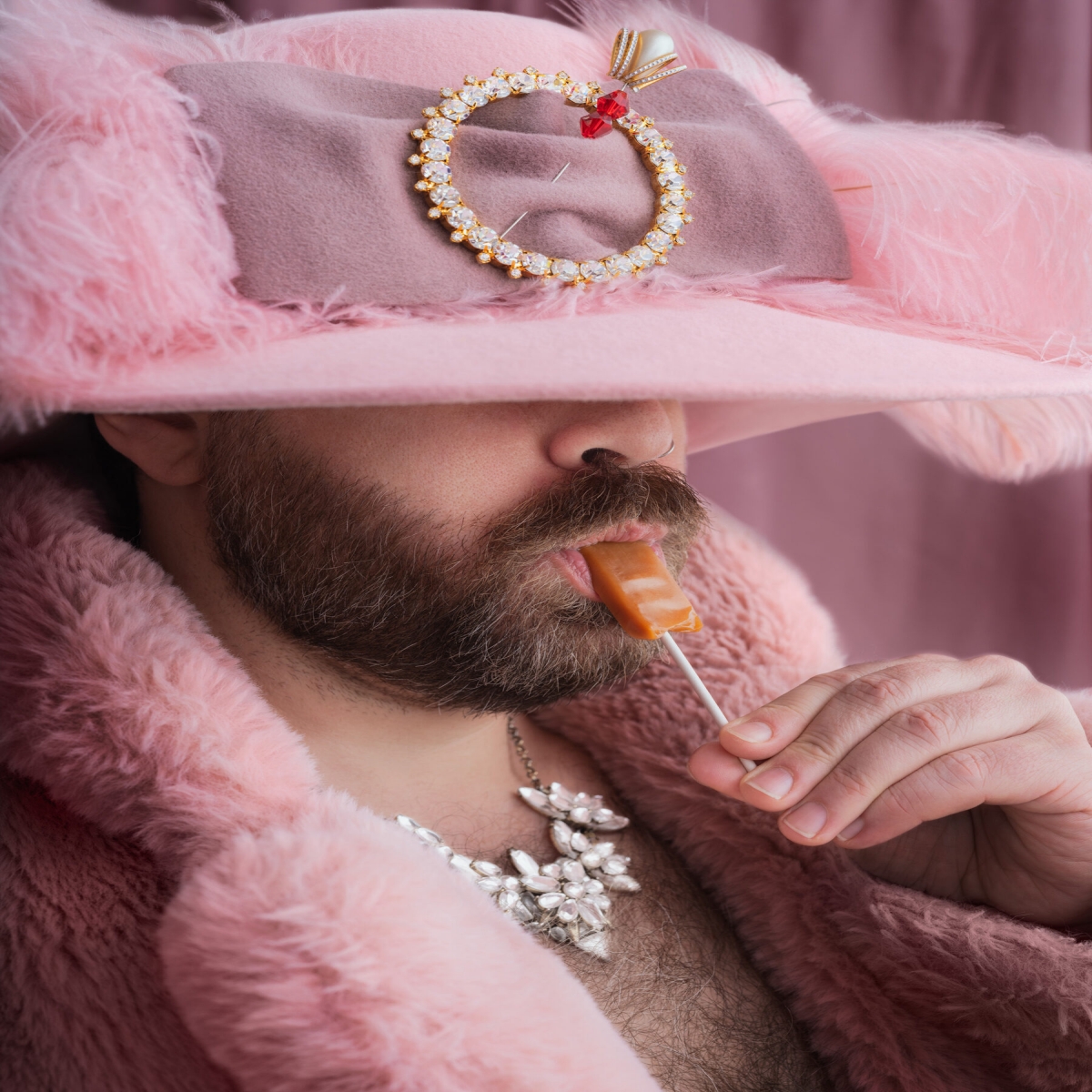 Sugar Daddy Exhibition Photo, featuring a masculine-presenting individual in a pink hat with a jeweled brooch, draped in furs, eating a lollipop