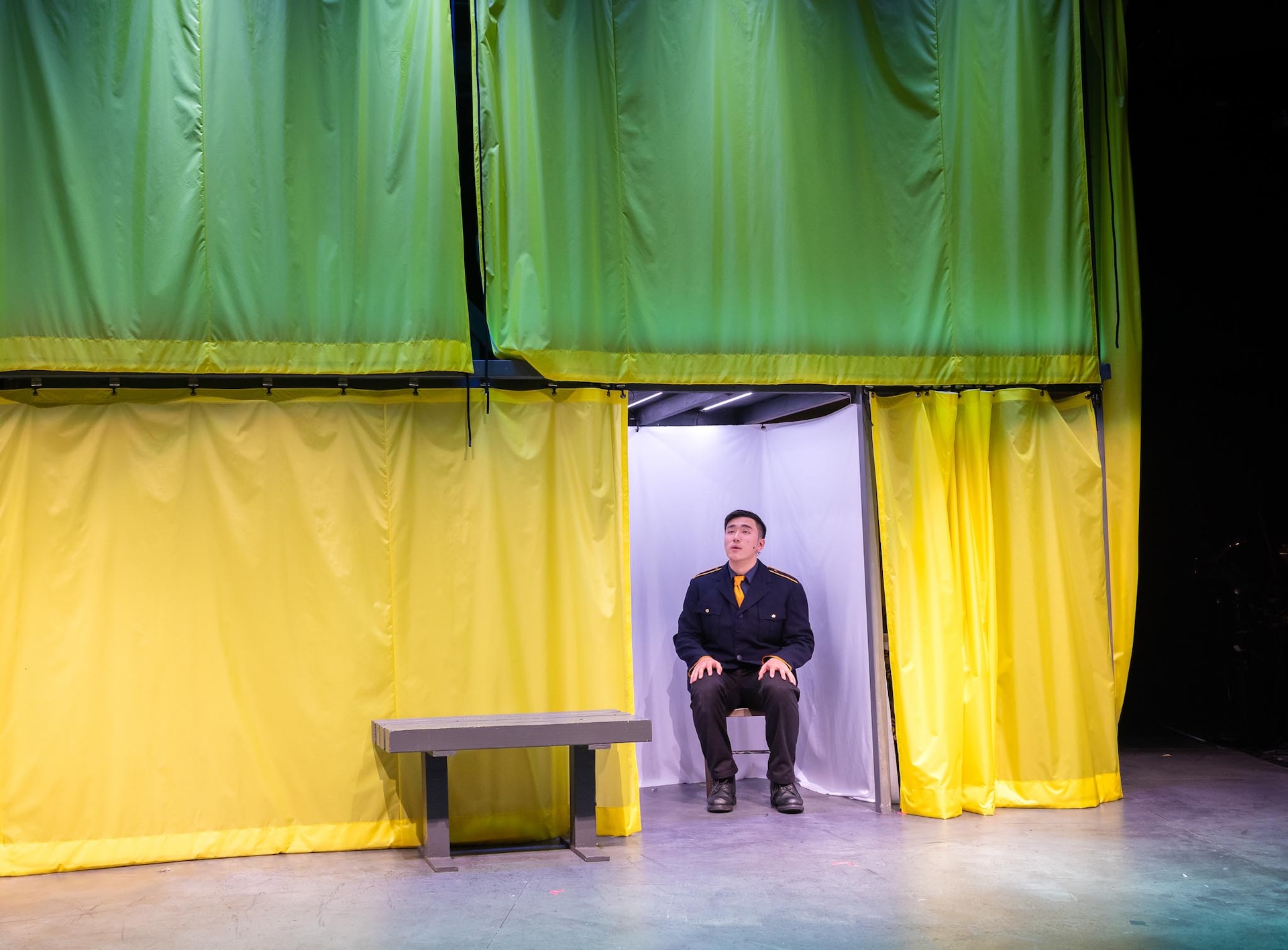 Image of set on stage. There is a wall with two coloured panels, green on top and yellow on the bottom. With a gap in the yellow section and you can see an actor sitting on a seat