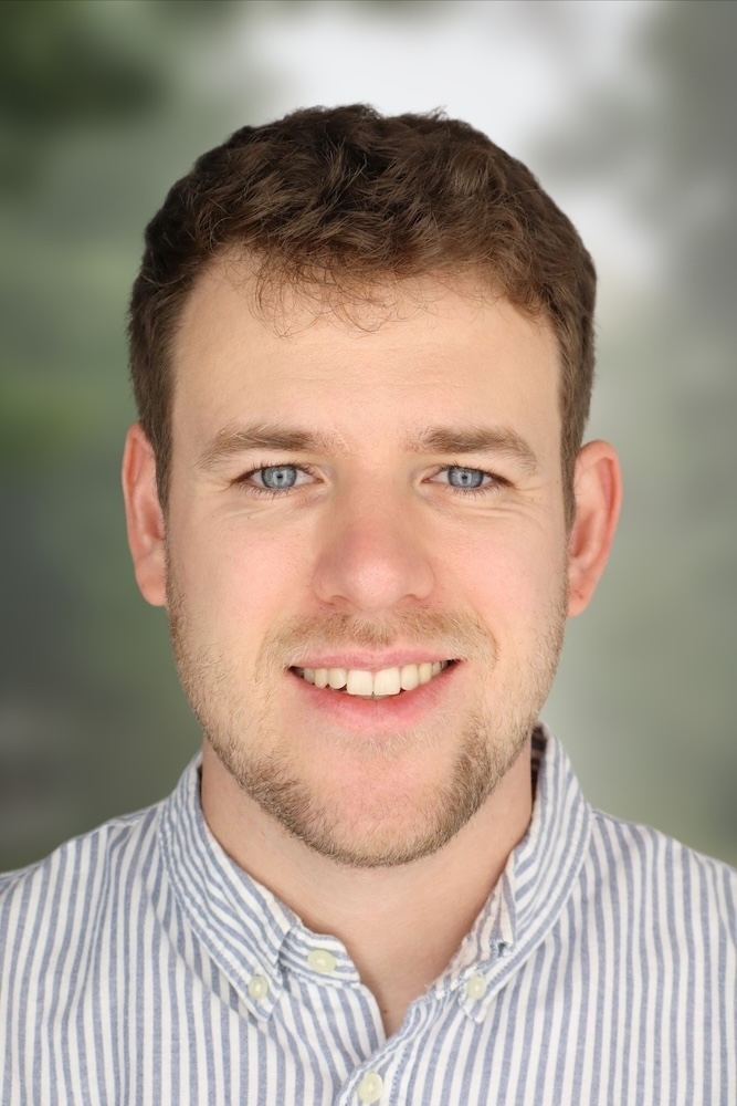 Young man with short brown hair and blue/grey eyes faces the camera smiling. He is wearing a pinstripe blue button down shirt.