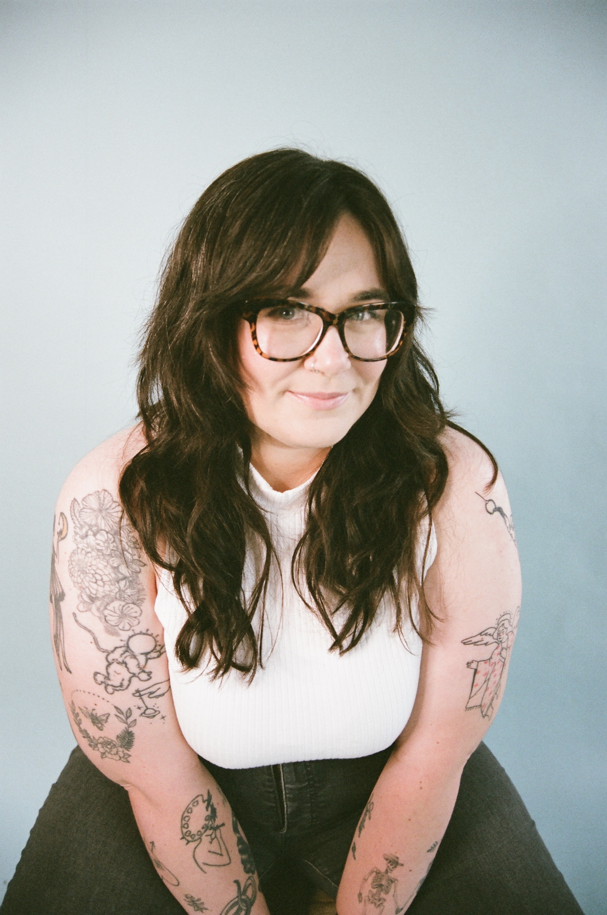 A woman smiles leaning forward into the camera. She has long wavy brown hair and tattoos on her arms.