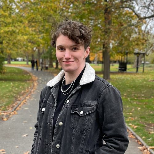 A woman with curly short brown hair is standing in the park smiling. She wearing a black denim jacket and black shirt with silver chains