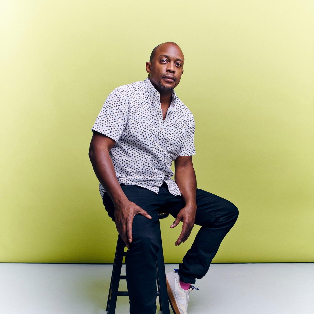 Gala honoree Hank Willis Thomas with a patterned shirt and blue pants sitting on a stool in front of a yellow wall. 