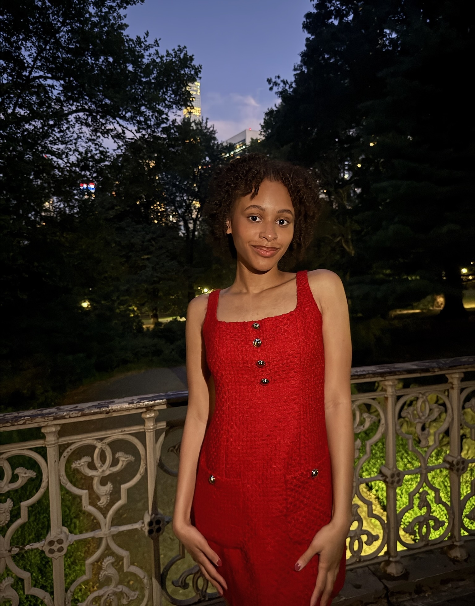 Girl with brown hair in red dress against a gate with trees in the background