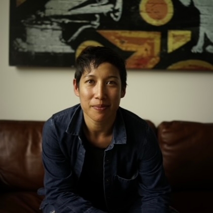 Professor Mariana sit on a leather couch wearing a jean jacket. Her dark hair is in a pixie cut. 