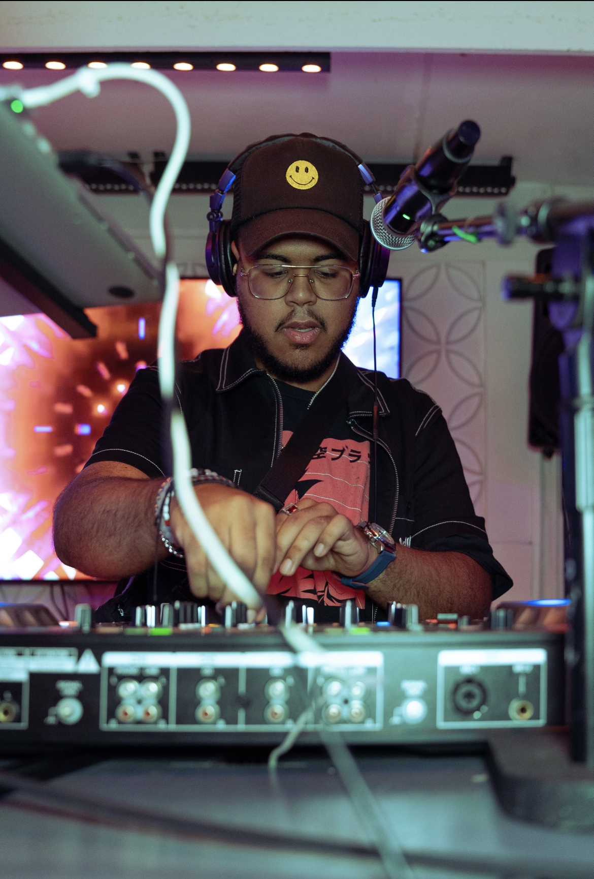 man in hat with smiley face on it works at DJ equipment. 