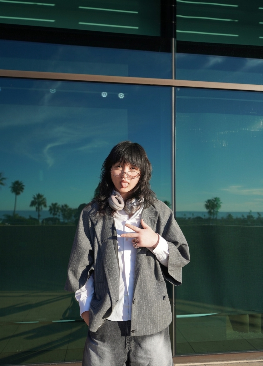 Student with mid-;length jacket up against a window, the window has reflections of palm trees