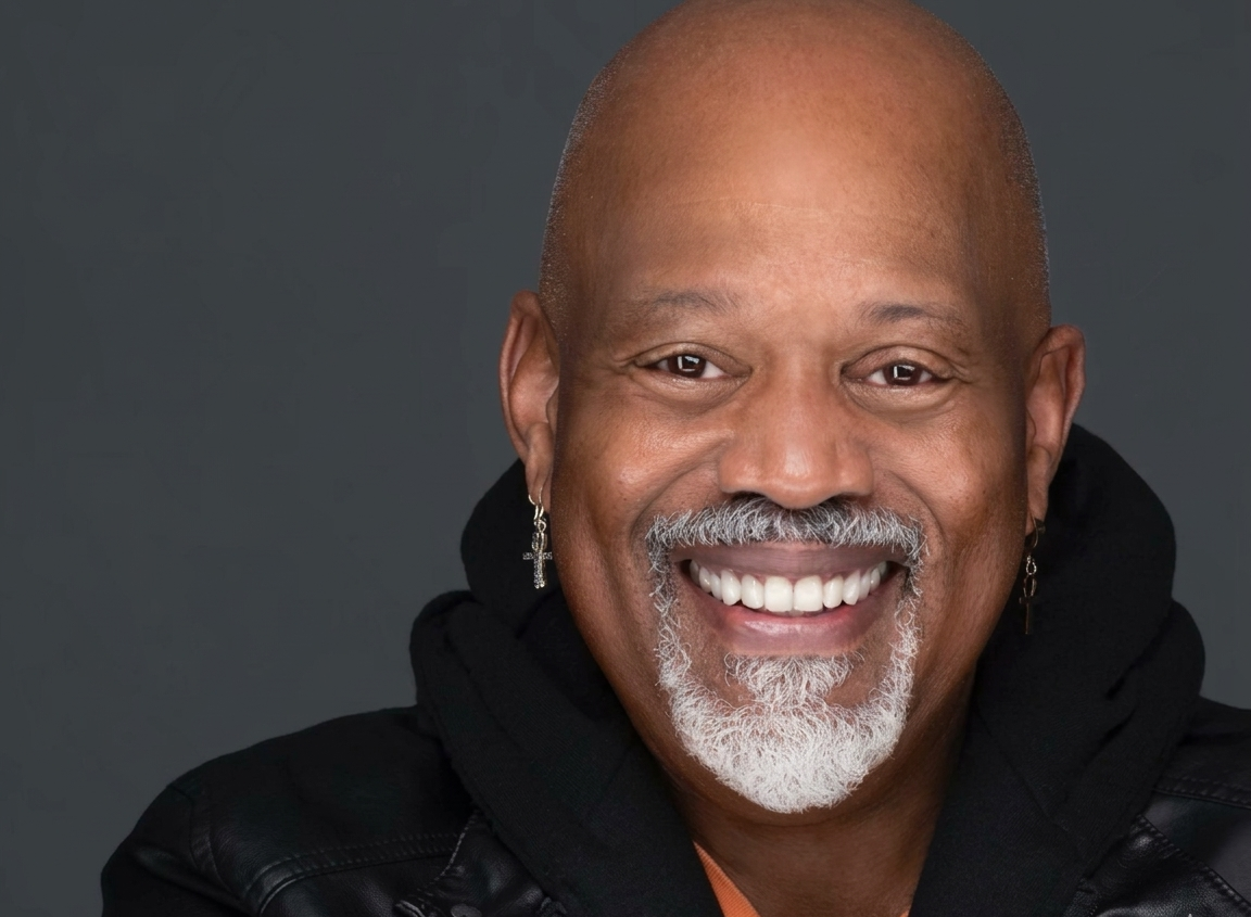 Smiling man with grey beard in a black jacket with orange underneath. 