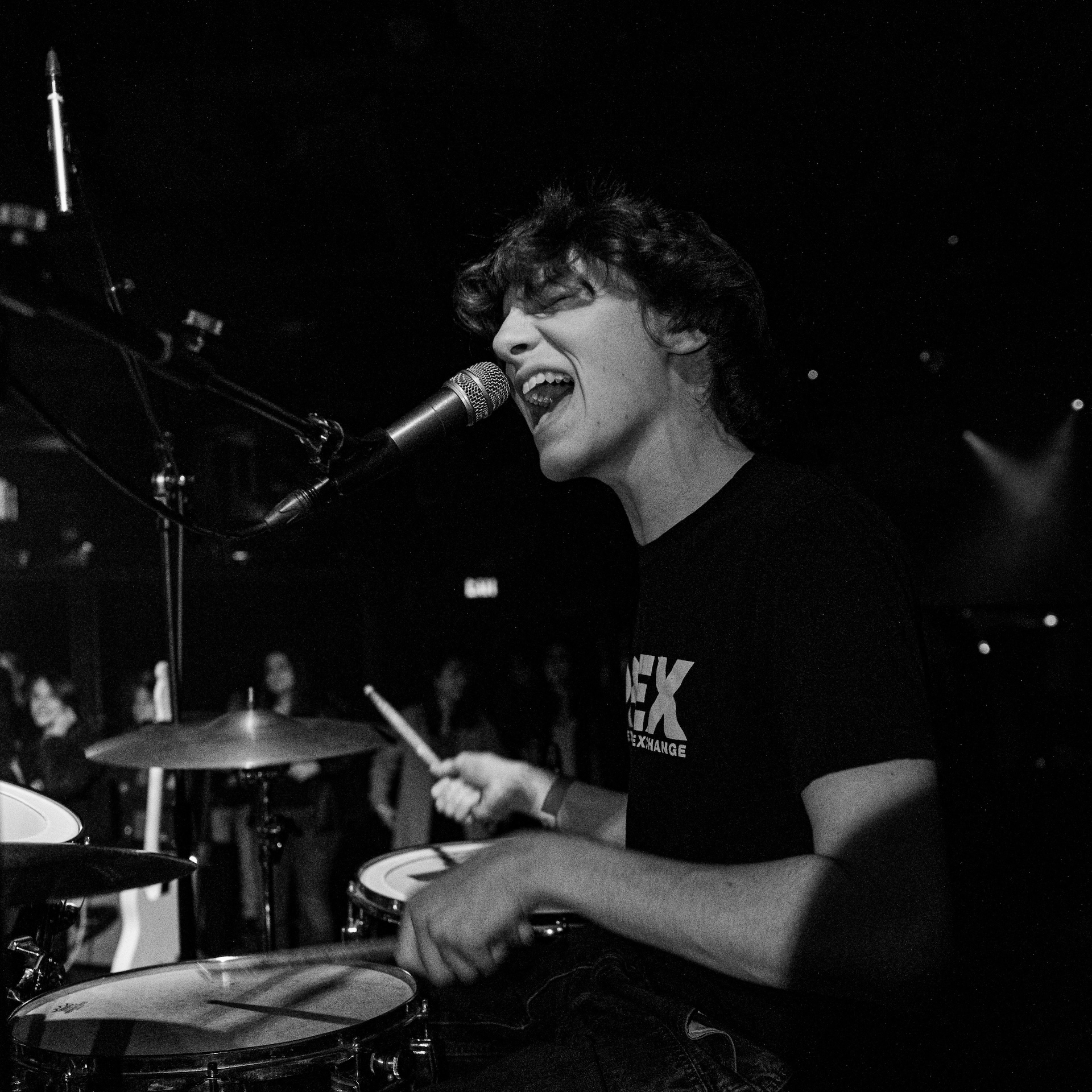 Student with short brown hair sitting at the drums singing into a microphone