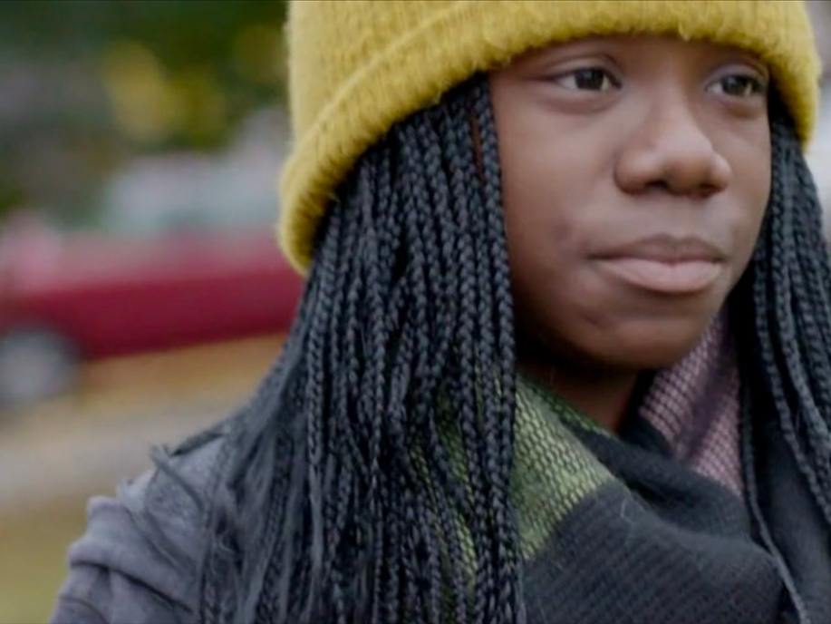 Film Still from Tween. Close up image of girl's face with mini-braids and yellow hat.