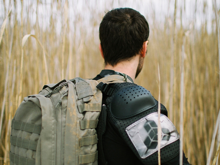 Film still from Hiraeth. Image of man from behind, he has an army backpack and shoulder pads on. 