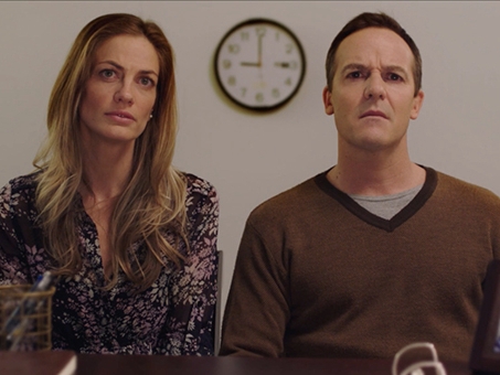 Film still from Leecher. Image of woman and man from waiste up, sitting with clock on wall behind them. 