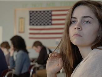 Film still from Northshore. Image of young woman in class with American flag in the background. 