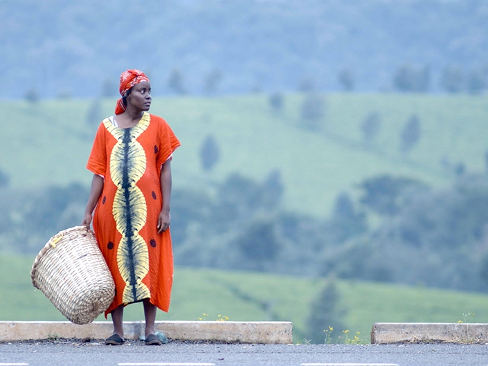Film Still from Chebet. Image of woman in orange and yellow dress with red scarce around her head waiting on a road with basket in her hand. 