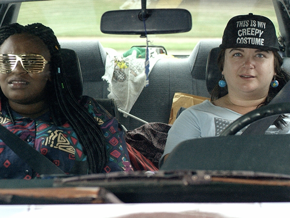 Film Still from DO NOT DISTURB. Image of two woman in the front seat of a car. One woman't cap reads, This is my creepy costume. 