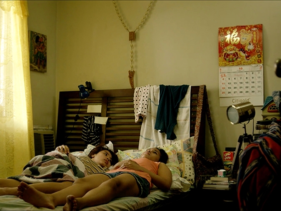Film Still from Ina Nyo. Image of two women sharing a bed in a messy room.