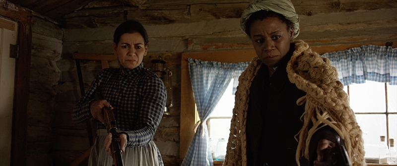 Two pioneer women standing in a cabin, one holding a shotgun.