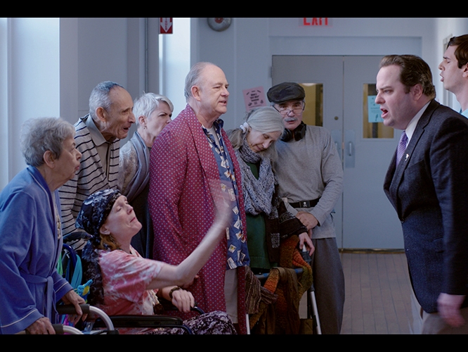 Film Still from Shady Pines. Image of woman in wheel chair surrounded by older people in confrontation with man in a suit.  