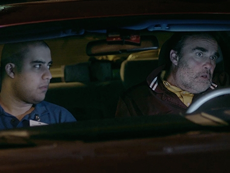 Film Still from The Stock Boy. Image of inside of a car from the front with two men, one driving, in the evening. 