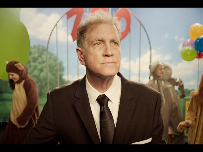 Film still from Willy. Image of man on TV set with blue sky backdrop and people in animal costumes behind him