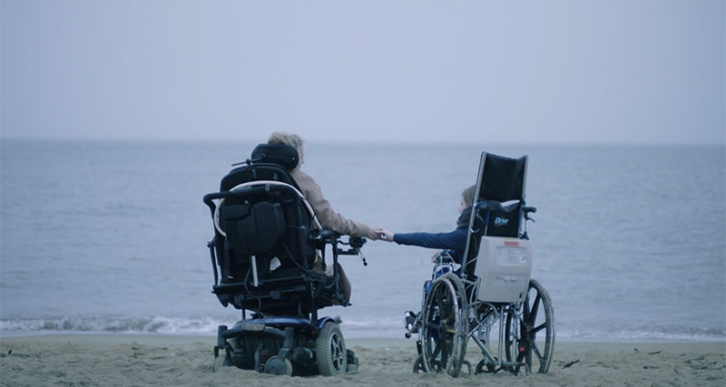 Two persons in wheelchairs, holding hands on a beach.