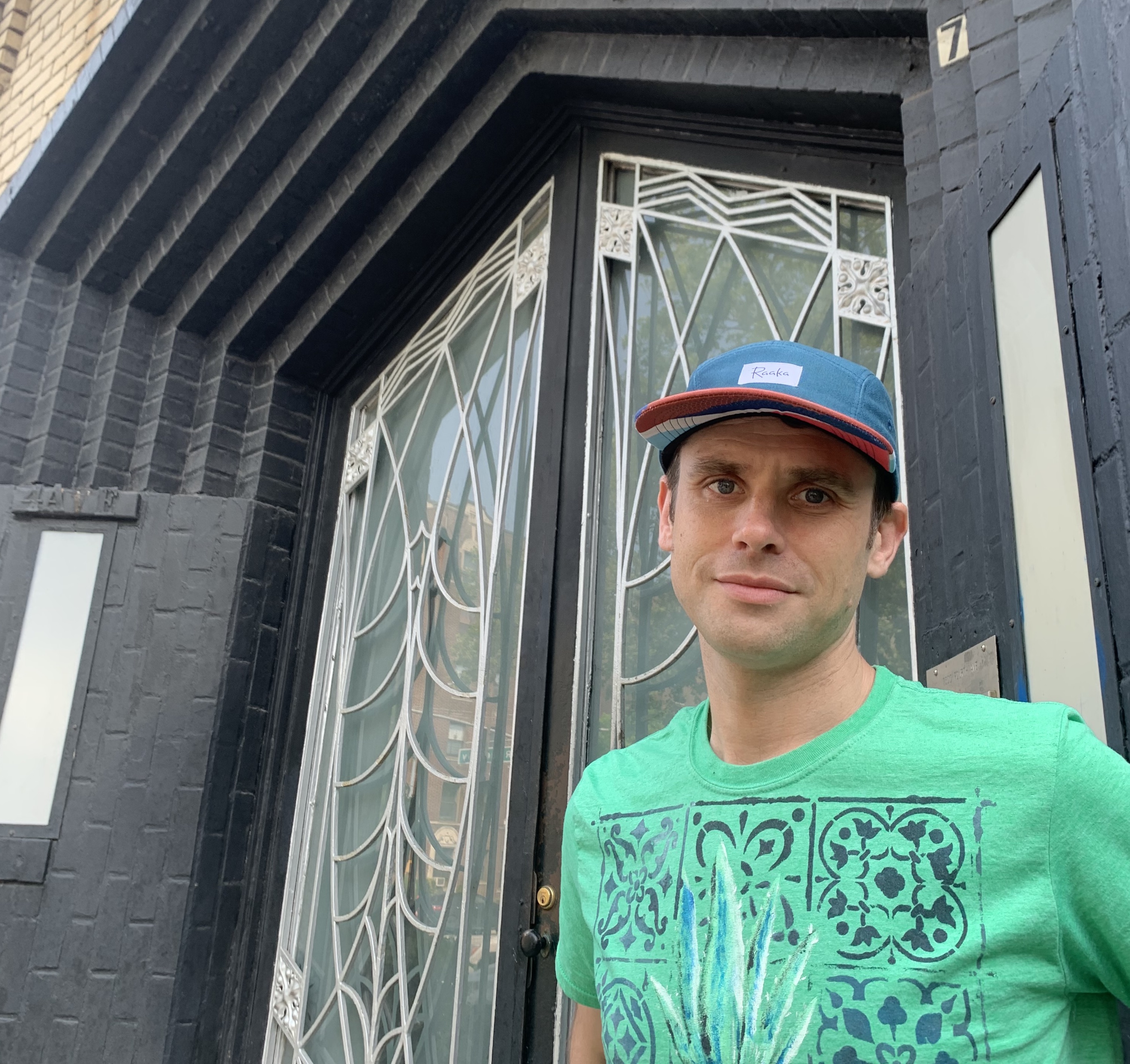 Greg wearing a green graphic shirt and blue hat standing in front of an elaborate door.
