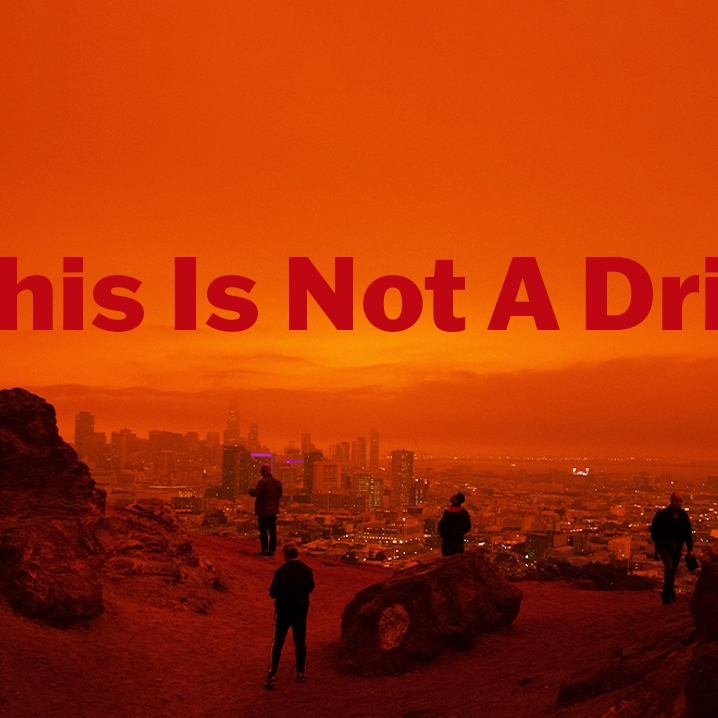an orange tinted image of people standing on a rock formation overlooking a city. The words: *This Is Not A Drill* are printed on top of the image
