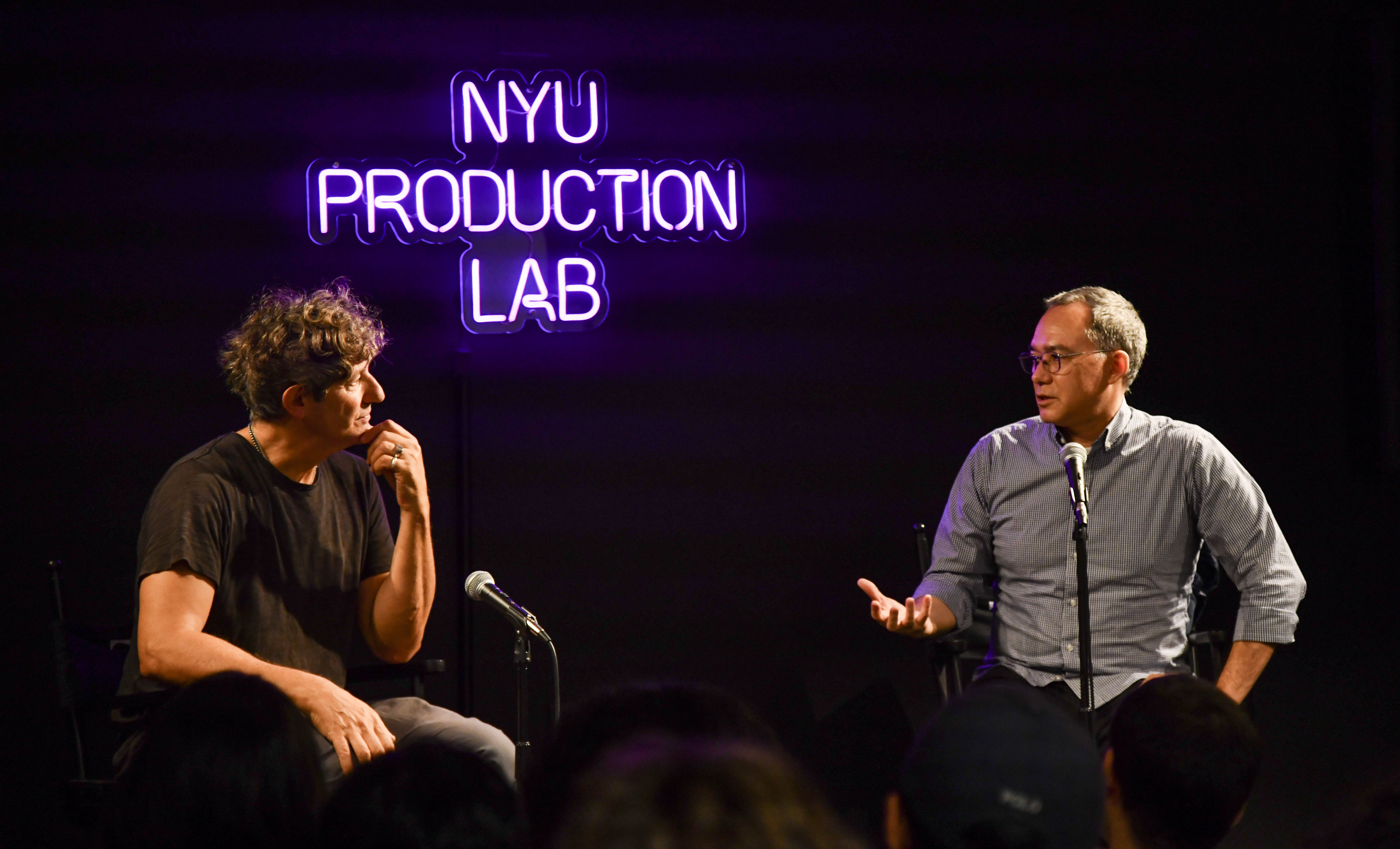 Filmmaker Jonathan Glazer sits with Grad Film Chair Andrew Okpeaha MacLean in front of a neon sign that reads "NYU Production Lab"