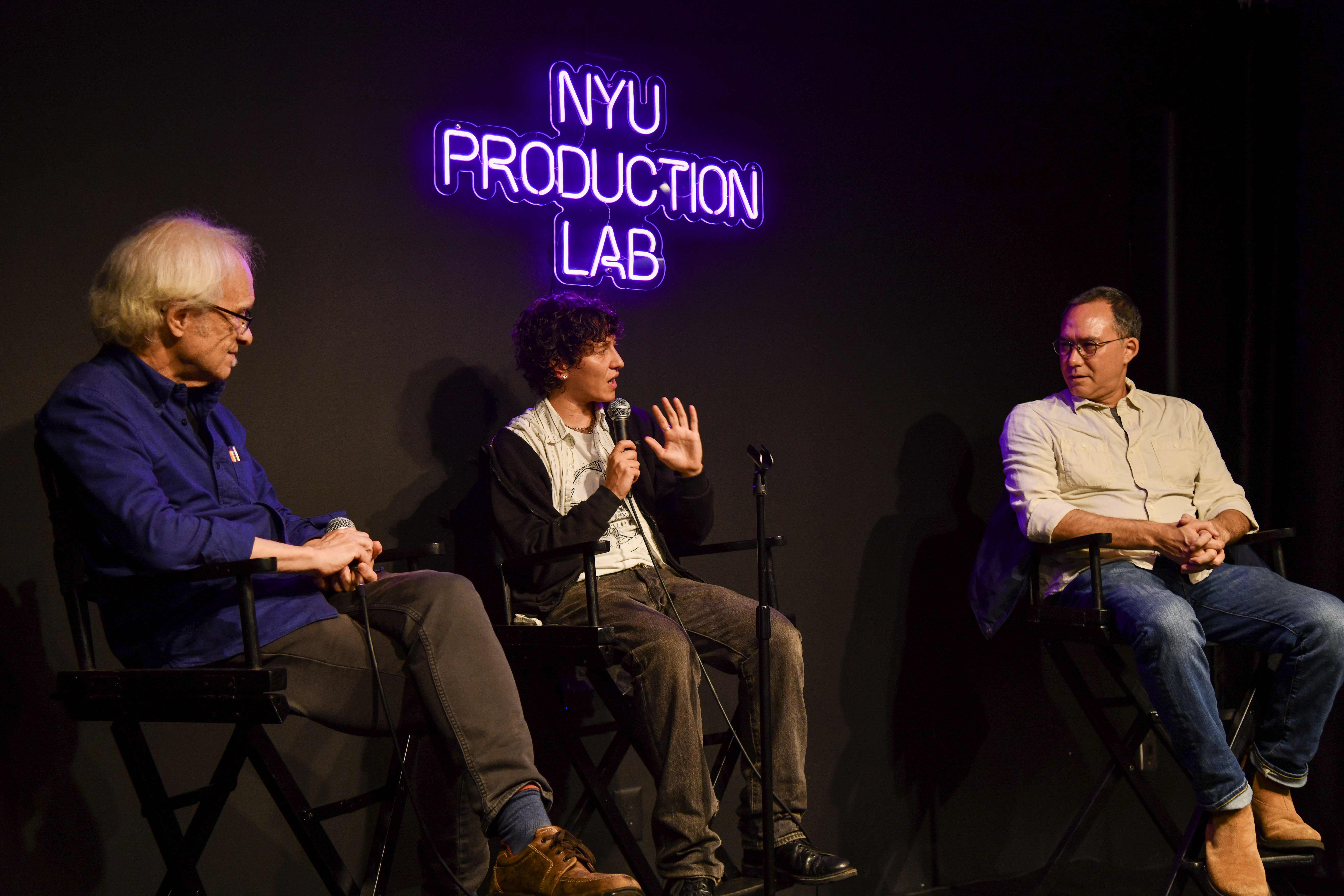 Filmmaker Mica Levi sits in conversation with Grad Film Chair Andrew Okpeaha MacLean and one other person in front of a neon sign that reads "NYU Production Lab"