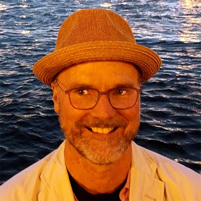 A man smiling wearing a hat with a lake behind him