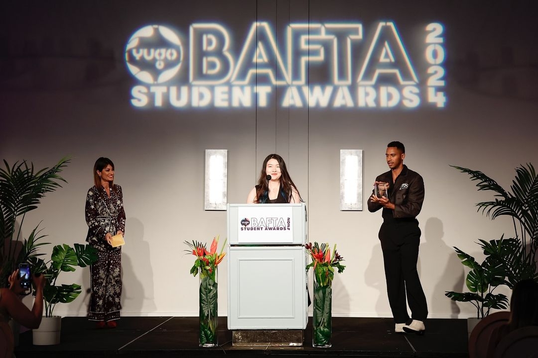 Three individuals stand on stage at the BAFTA Student Awards 2024. A woman in a patterned dress is on the left, smiling and holding a paper. In the center, a woman is speaking at a podium with a BAFTA Student Awards logo. On the right, a man in a dark shirt and pants is holding a clear award trophy.
