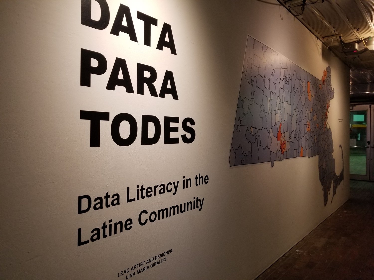 Text on wall that says "Data Para Todes: Data Literacy in the Latine Community. Lead artist and designer Lina Maria Giraldo" and a picture of a map with borders.