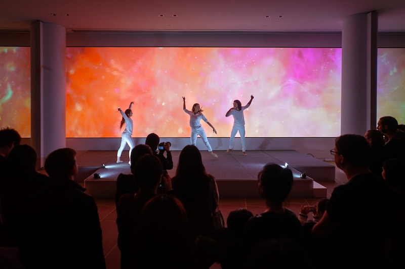 Dancers dance on stage with colorful screen behind them